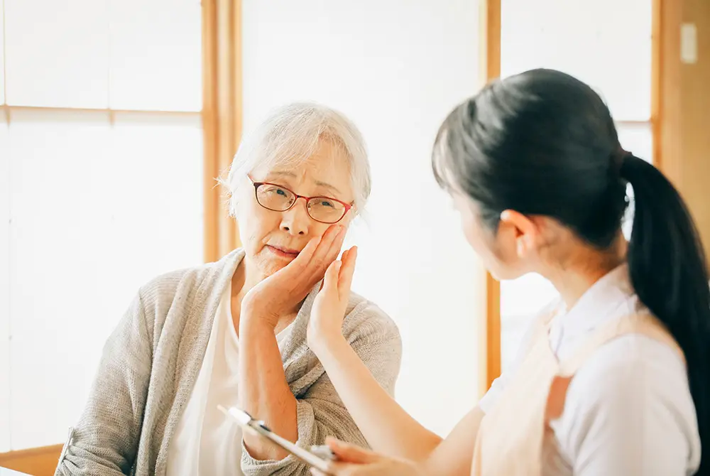 頬を押さえながら痛みを訴える高齢女性と話を聞く医療従事者。