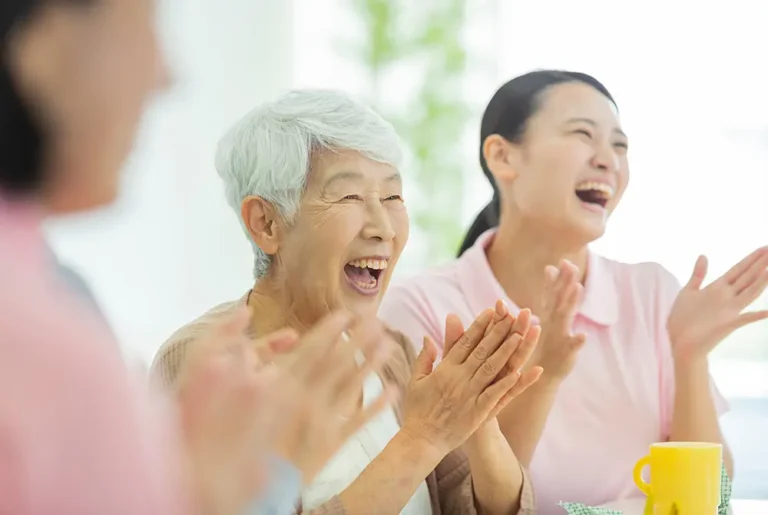 高齢女性と介護スタッフが笑顔で拍手している様子。