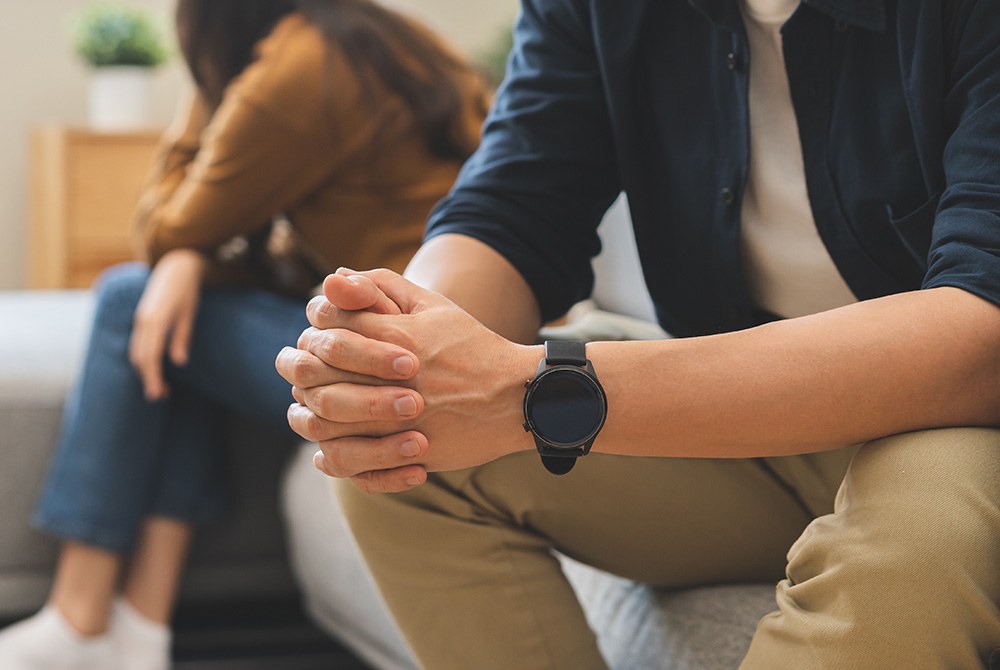 ソファに座り悩んだ表情で手を組む男性と、後ろで距離を置く女性