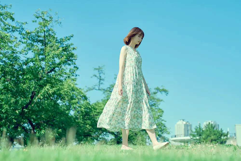 晴れた公園で、淡い色のワンピースを着た女性が芝生の上を歩く様子。