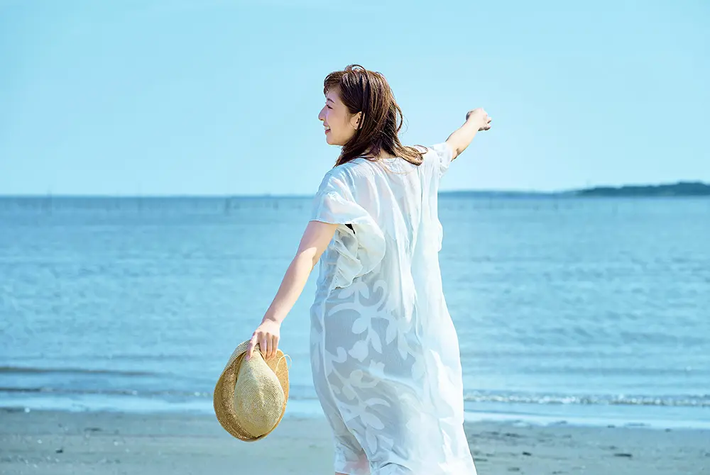 海辺で帽子を手に持ち、白い服を着て歩く女性。