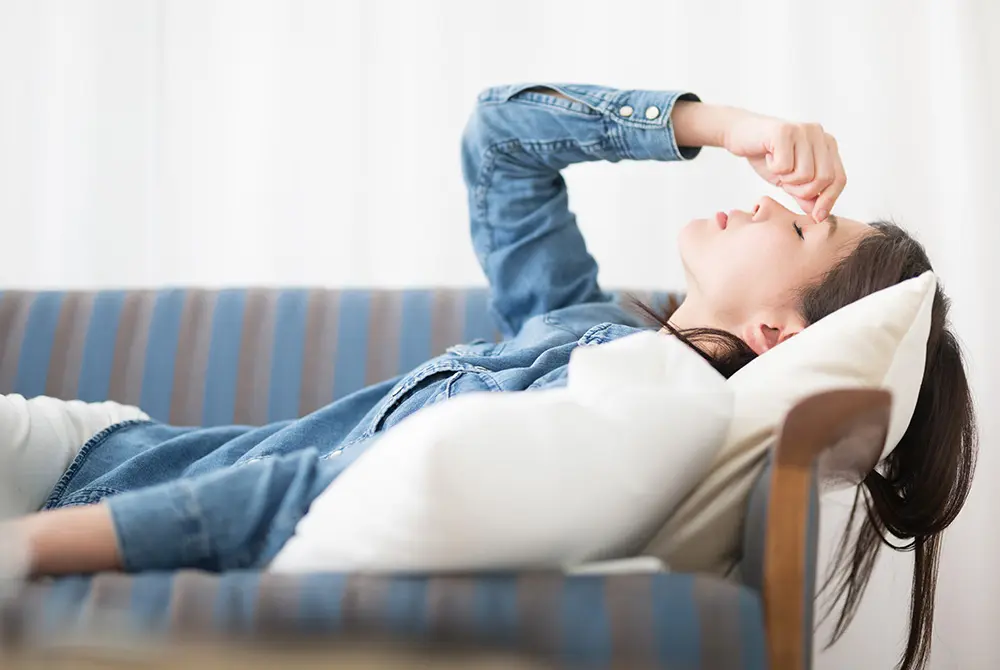 ソファに横になり、鼻のあたりに手を当てる女性。体調不良や疲労を示すイメージ。