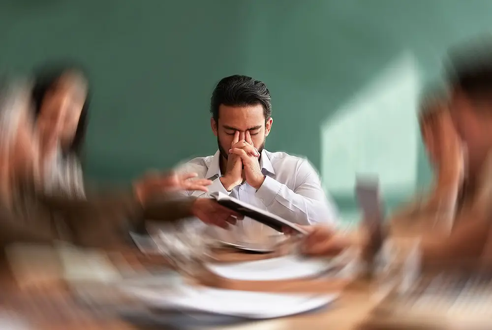 会議中に目を閉じ、鼻の付け根を押さえる男性。ストレスや不調を示すイメージ。
