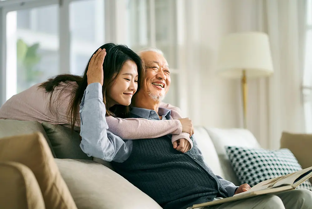 ソファに座る高齢男性に、若い女性が後ろから寄り添っている様子。