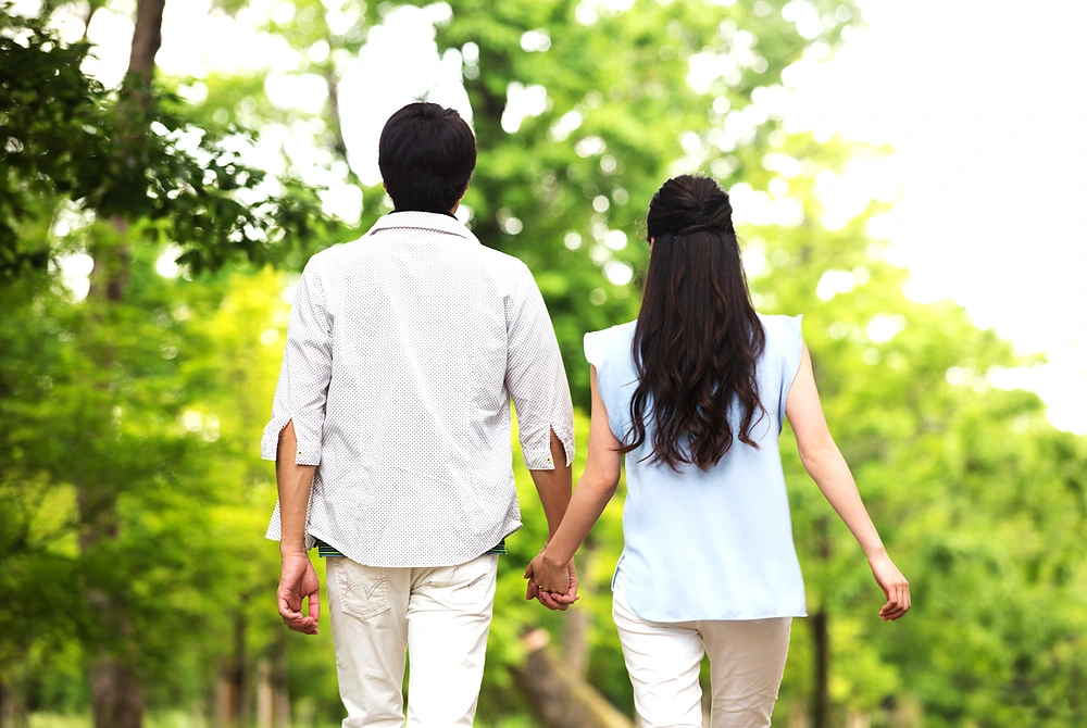 緑の多い道を手をつないで歩く男女の後ろ姿。