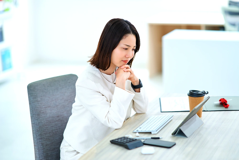 机の前で端末を見ながら、自分の体調や生活習慣について考える女性。