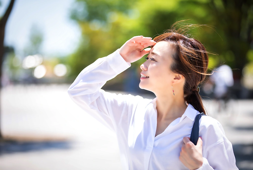 強い日差しの中で手を額にかざし、まぶしそうに空を見る女性。