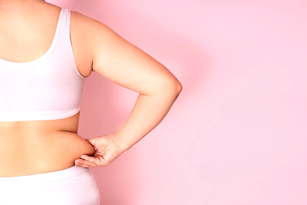 ピンクの背景の前で、腹部の脂肪を手でつまむ人の上半身。