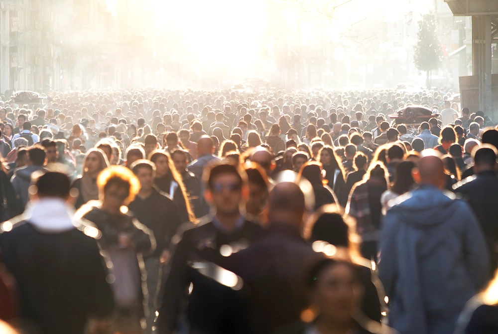 強い日差しの中、都市部の通りを多くの人が行き交う様子。