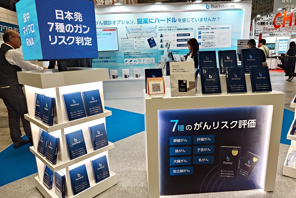ブルーインダストリーズの「bluedrop」展示ブース。尿中マイクロRNAによる7種類のがんリスク評価を紹介するパネルと製品資料が並ぶ。