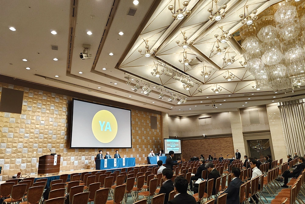 第54回日本磁気共鳴医学会大会の会場内。大型スクリーンの前に複数の登壇者が並び、参加者が着席して講演や討論を聴いている。