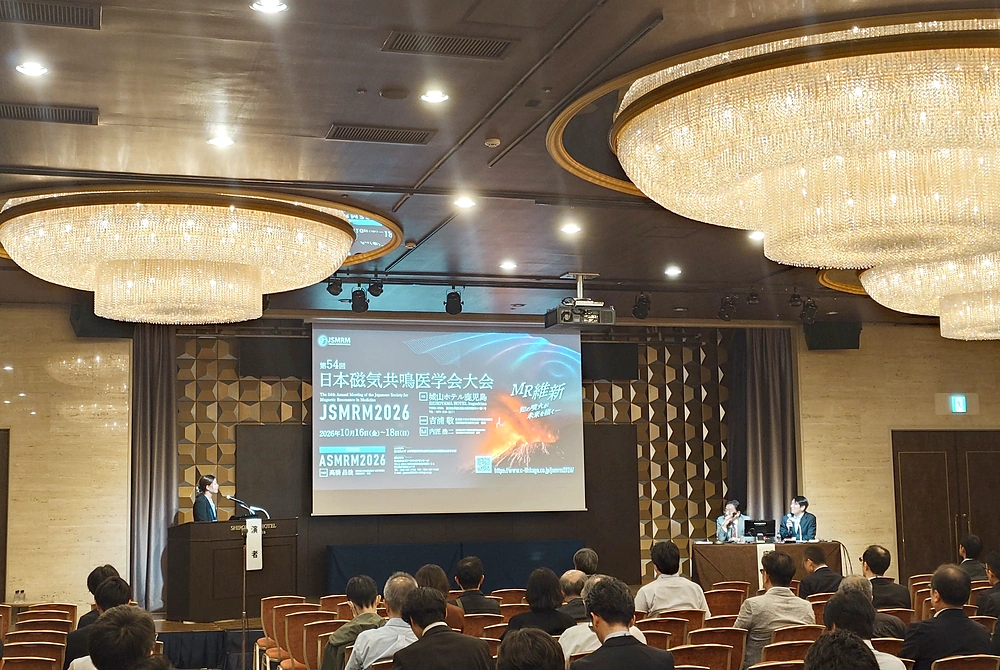 第54回日本磁気共鳴医学会大会のシンポジウム会場。演台で講演する登壇者と座長、講演を聴く参加者の様子。