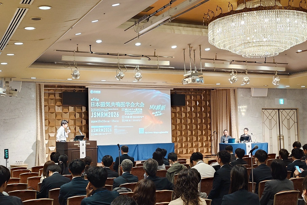 第54回日本磁気共鳴医学会大会のシンポジウム会場。演台で講演する登壇者と座長、講演を聴く参加者の様子。