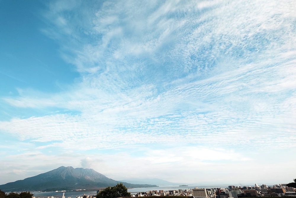 鹿児島市街地から望む桜島。手前に市街地と錦江湾が広がり、青空には薄い雲が一面に広がっている。