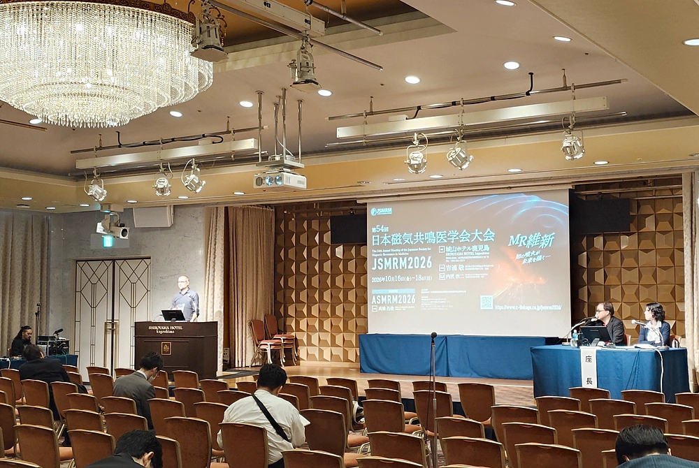 第54回日本磁気共鳴医学会大会の講演会場。演台で講演する登壇者と、座長席で話を聞く2人の座長、参加者の様子。