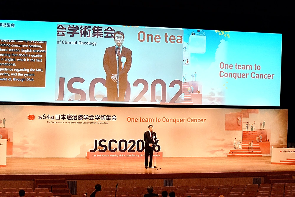 第64回日本癌治療学会学術集会の開会式。大型スクリーンに登壇者の姿と大会テーマが映し出され、ステージ中央であいさつが行われている。