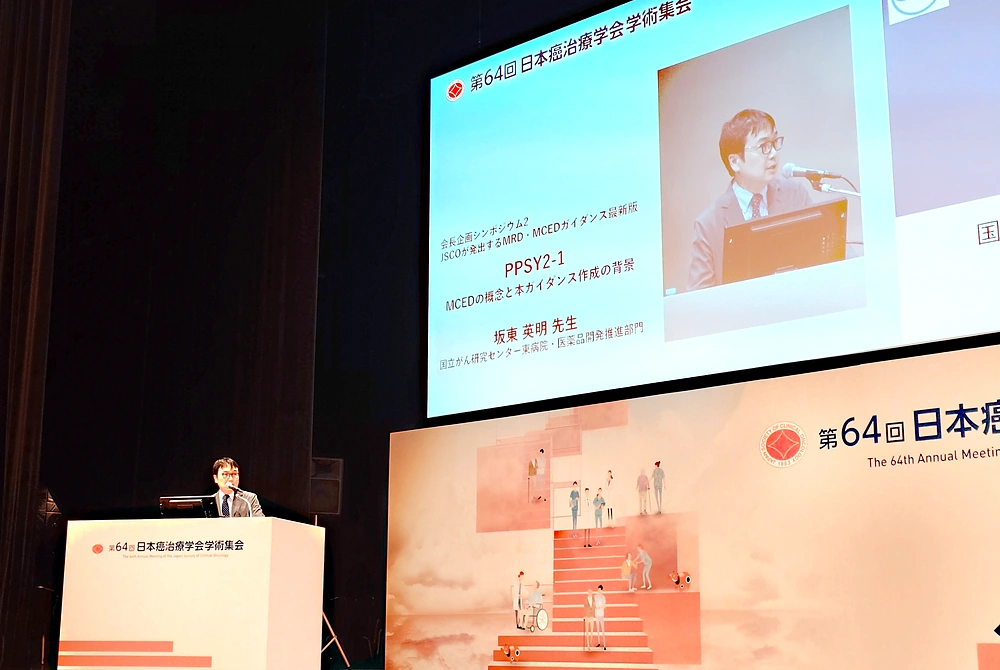 第64回日本癌治療学会学術集会の講演会場。大型スクリーンには「MCEDの概念と本ガイダンス作成の背景」と表示され、坂東英明氏の講演が紹介されている。