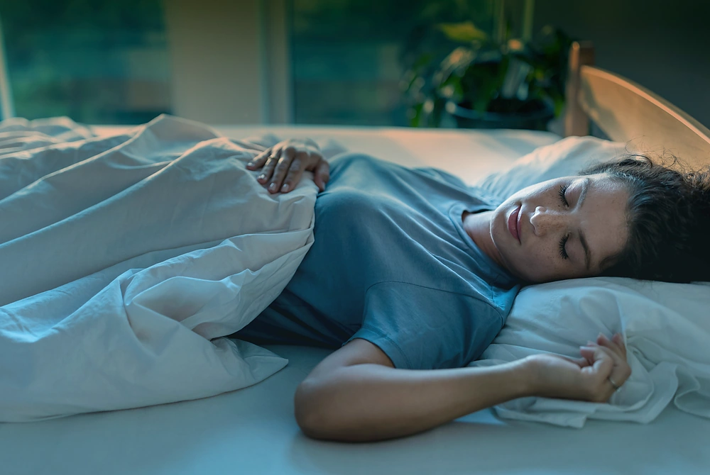 薄暗い寝室で、掛け布団をかけて横向きに眠る女性。枕に頭を乗せ、目を閉じている。